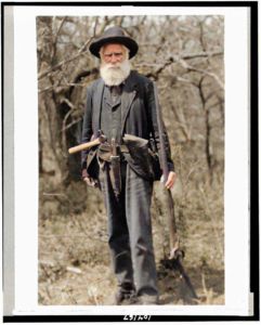 Old man with long white beard and hat standing holding a rifle and with a hatchet tucked into his belt and standing outside