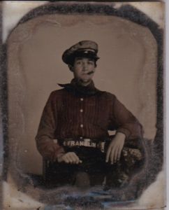 seated young man with a neck bandana, cigar, billed cap, dark trousers, loose-fitting shirt, and a belt that has on it 4 Franklin 4