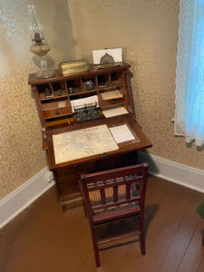 An upright wooden writing desk with various built-in small shelves and drawers with an oil lamp and a letter holder on top of the desk and a wooden chair for the desk