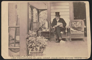 Bearded old man seating on an outdoor porch while reading a newspaper and wearing a top hat, round-framed spectacles, light-colored silk scarf, and overcoat with man in seated in the shadow of the doorway nearby.