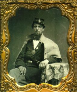 A soldier, in a seated pose with one leg crossed over the knee of the other, having facial hair of the goatee style and wearing a uniform including a bowtie and kepi cap with a metal letter 