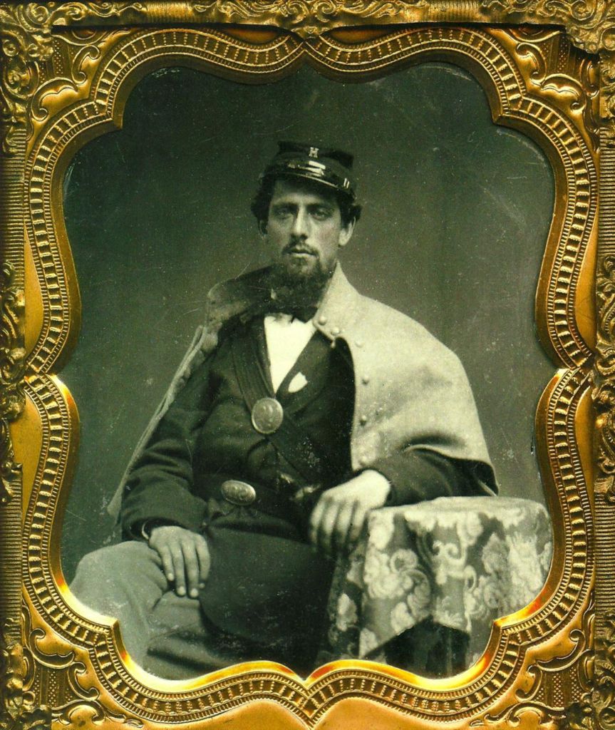 A soldier, in a seated pose with one leg crossed over the knee of the other, having facial hair of the goatee style and wearing a uniform including a bowtie and kepi cap with a metal letter "H" pinned to its front