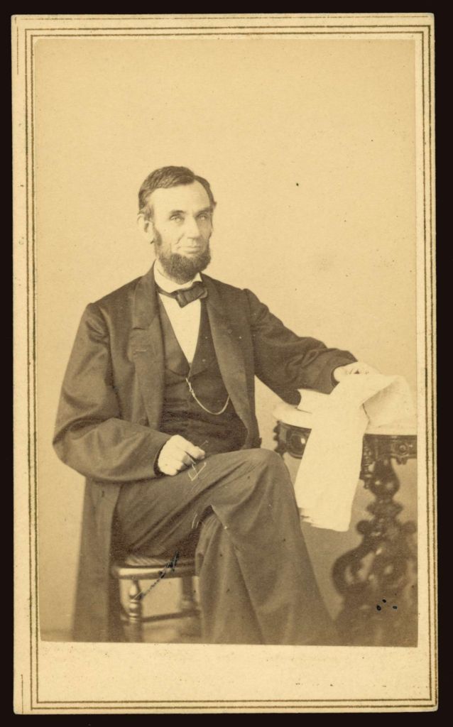President Abraham Lincoln with hair parted on his left and with a beard without a moustache (which is known as a chin curtain) poses seated with legs crossed in a dark suit, white shirt, dark bowtie, dark waist coast with the chain of a pocket watch in a waist coat pocket visible. He his seated next to a small, round dark-wood table with a white marble top on it and resting his left arm on that table, in his left hand is a newspaper; in his right hand that is resting on his right thigh is a pair of wire-frame reading glasses.