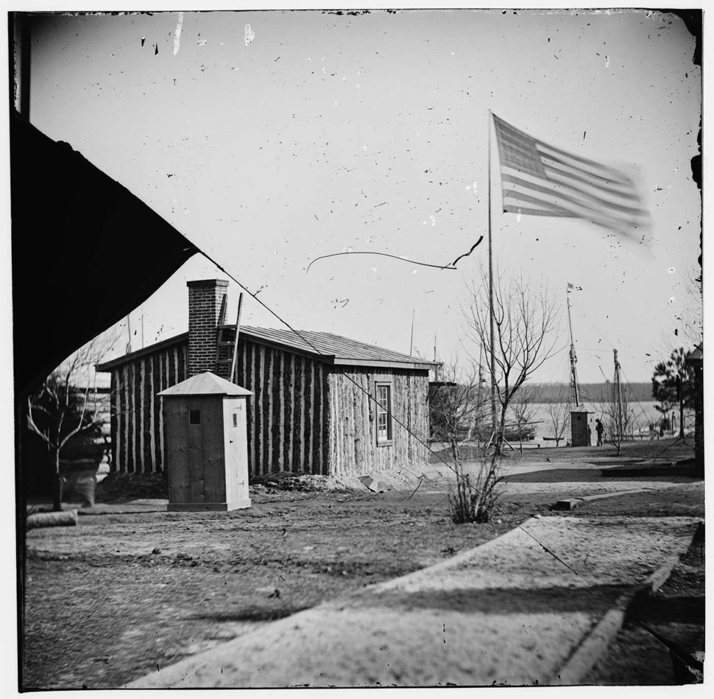 The view of the back of a small, one-story, house-like structure of vertical wooden timbers, a brick chimney, and a slightly sloped shingled roof, and it is situated on rough, grassless ground with a United States flag raised on a flagpole by it and flying vigorously in the breeze 