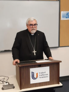 clergyman with white hair, white goatee, large black spectacles, standing behind a portable tabletop lectern and wearing black clothing including a black shirt with a black band collar and white clerical tab visible in the center of the collar and a black jacket and a large silver unadorned pectoral cross suspended on a very long silver necklace chain, the cross designating status as a bishop or similar high office within the church; on the lectern is a sign reading 