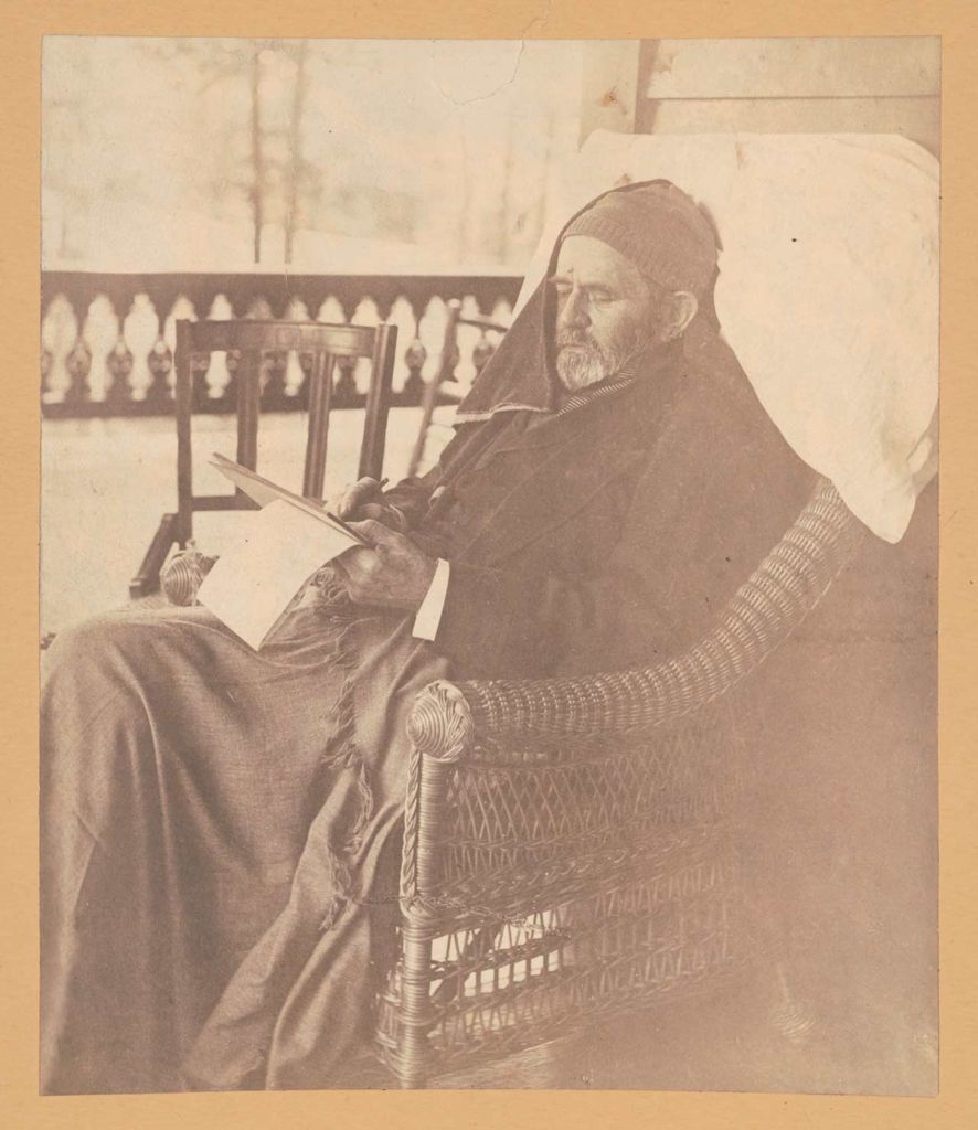 Ulysses S. Grant, three-quarter length portrait, bearded, wearing a cap, seated in rattan chair, a blanket over his lap, a pillow behind his head, a towel draped very partially over the side of his face away from the viewer, writing his memoirs, at Mount McGregor, Wilton, New York, near Saratoga Springs.