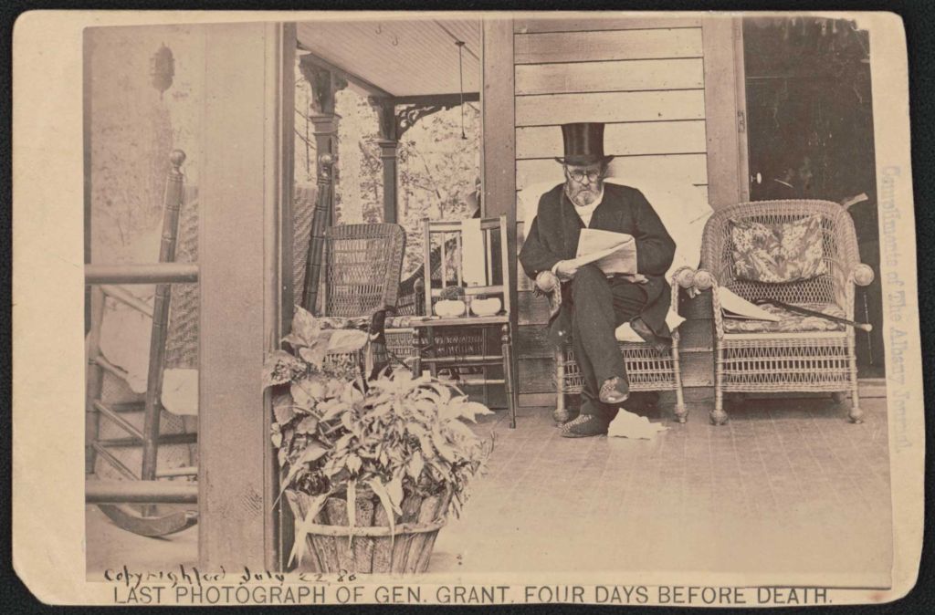 Bearded old man seating on an outdoor porch while reading a newspaper and wearing a top hat, round-framed spectacles, light-colored silk scarf, and overcoat with man in seated in the shadow of the doorway nearby