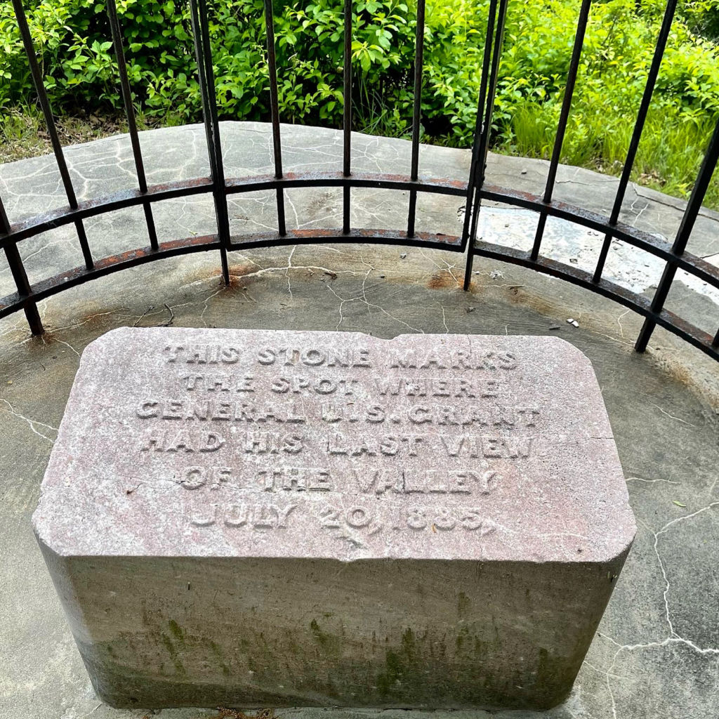 A gray, cube-like stone marker about two feet high by 2.5 feet wide set in a concrete base and encircled within a round metal fence painted black and on the top surface of the stone in relief are carved the words: This stone marks the spot where General U.S. Grant had his last view of the valley, July 20, 1885