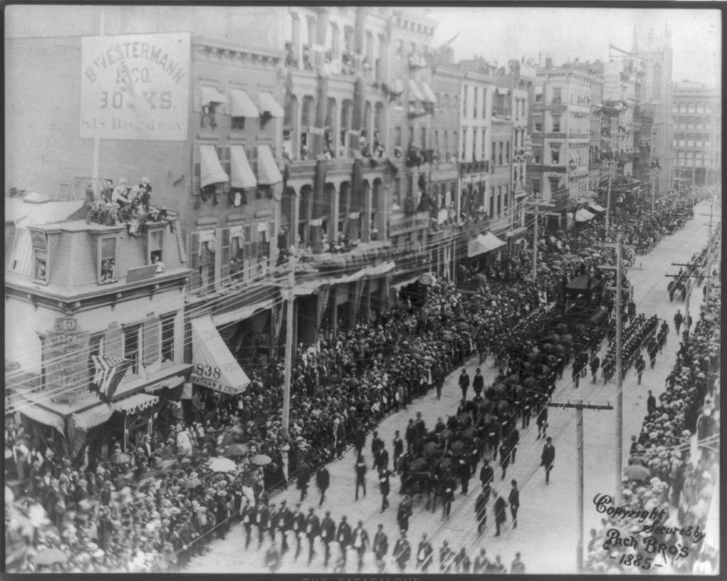 A huge crowd of spectators lining the streets, watching out of windows, and watching from rooftops as the funeral procession of President Ulysses S. Grant passes by in the middle of the street and including numerous men in dark clothes in orderly rows and Grant's catafalque being pulled by several teams of horses.